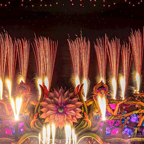 A vibrant, large-scale music festival at night with colorful stage decorations, fireworks, and a crowd. "Orlando" is spelled out in the sky above the event.