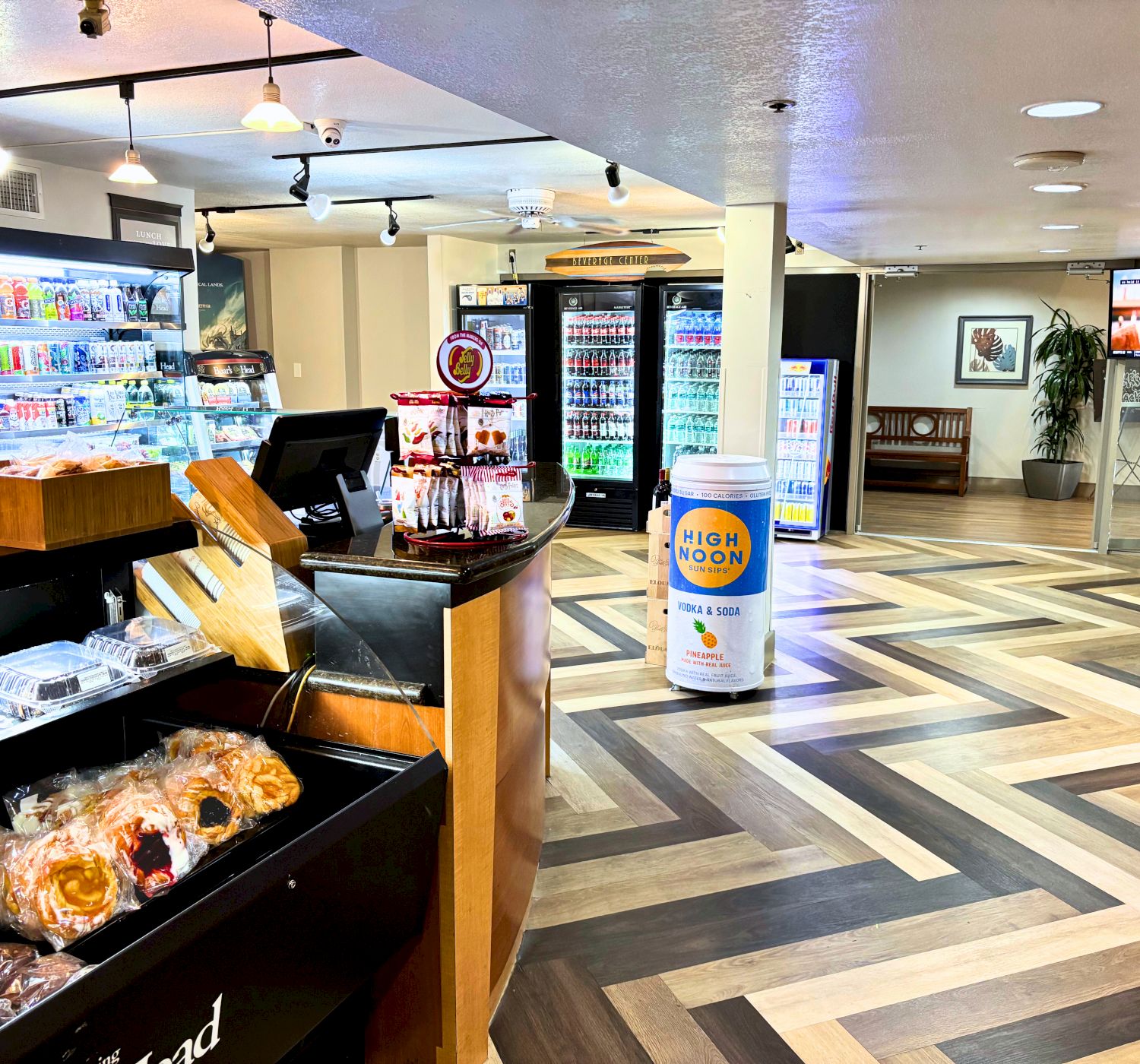 A convenience store with display cases of beverages and snacks, a coffee machine, and assorted products on shelves in a patterned floor setting.