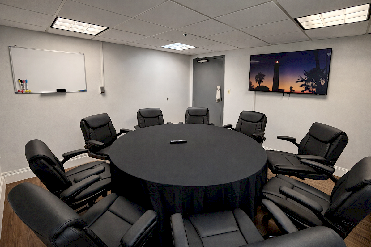 A conference room with a round table, black chairs, a whiteboard, and a wall-mounted screen showing a scenic image.
