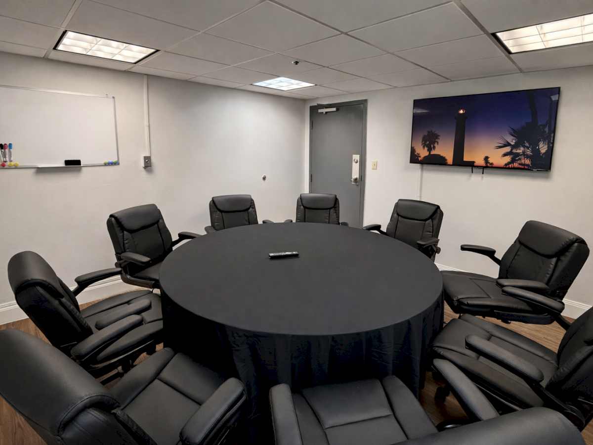 A conference room with a round table, black chairs, a whiteboard, and a wall-mounted screen showing a scenic image.