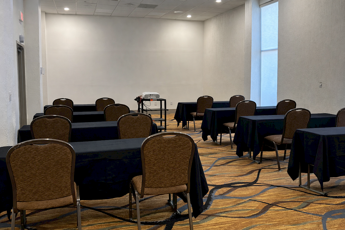 The image shows a conference room with several tables and chairs arranged in rows, and a projector on a stand at the front.