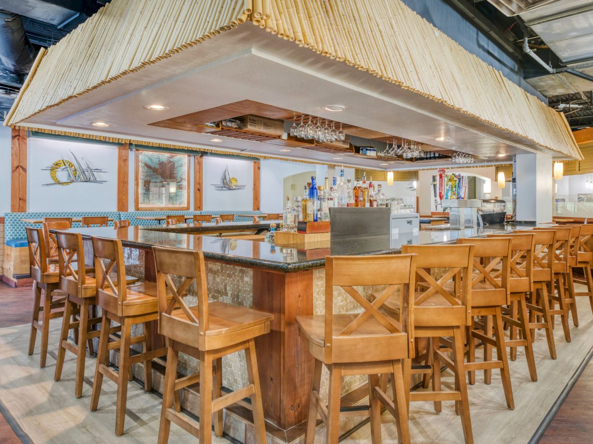 This image shows an empty bar with a wooden counter and chairs, surrounded by various bottles and glasses on display.