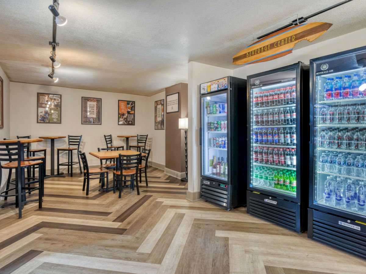 The image shows a small seating area with tables, chairs, and beverage coolers filled with drinks, set in a room with wood flooring.