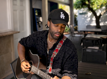 A person wearing a cap plays an acoustic guitar on a patio, surrounded by chairs and greenery, under string lights.